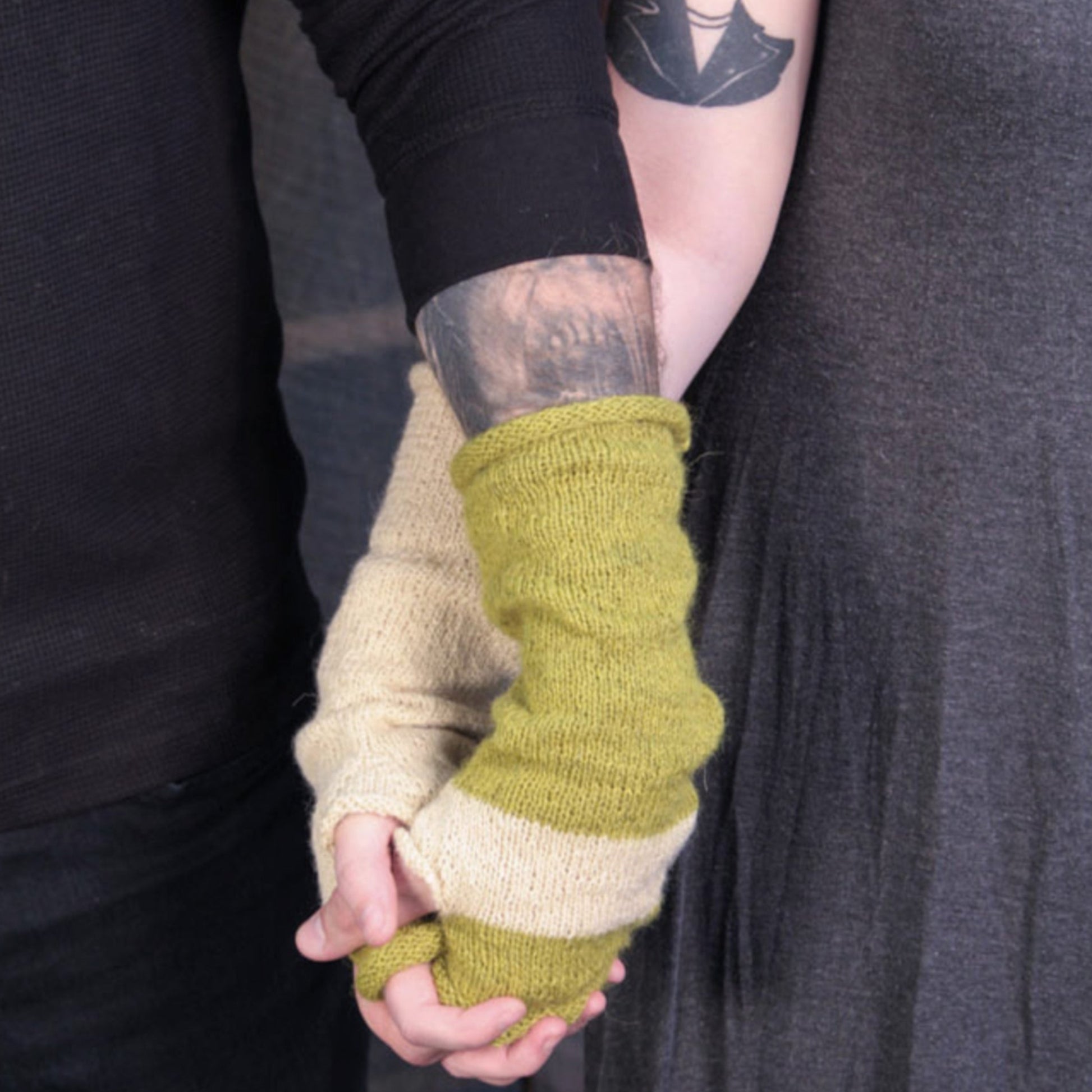 Two individuals holding hands, one wearing the "Whole Wide World" fingerless mitts by Halcyon Yarn, which feature a green and cream mixed design resembling slouchy mitts, and the other in a black long sleeve. The person in the slouchy fingerless mitts has a visible tattoo on their arm. Both appear to be dressed casually, with the background dark and out of focus.