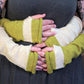 A person with tattooed arms wearing a dark gray dress is shown wearing the Whole Wide World Fingerless Mitts by Halcyon Yarn in a green and beige color block made of baby alpaca. The person's hands, adorned with these fingerless mitts, are clasped around their waist, one hand over the other.