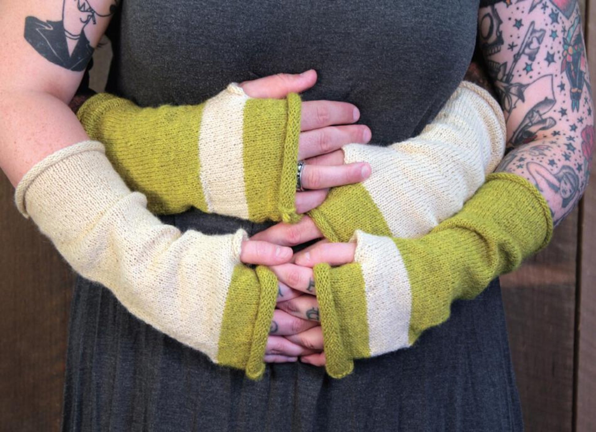 A person with tattooed arms wearing a dark gray dress is shown wearing the Whole Wide World Fingerless Mitts by Halcyon Yarn in a green and beige color block made of baby alpaca. The person's hands, adorned with these fingerless mitts, are clasped around their waist, one hand over the other.