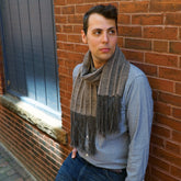 A person wearing a light blue dotted shirt and Halcyon Yarn's "Chain Reaction" woven scarf with an intricate weaving pattern leans against a brick wall in an alley. The person glances to the side with a thoughtful expression. They have short, dark hair and are surrounded by a peaceful, sunlit urban setting.