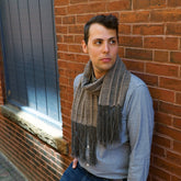 A person with short hair is leaning against a brick wall outside, wearing a light blue button-down shirt and a Chain Reaction woven scarf from Halcyon Yarn. They are looking to the side with a thoughtful expression.