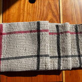 A Classic Linen Dishcloth from the Halcyon Yarn Weaving Kit (Rigid Heddle), featuring a textured beige fabric with black and red stripes, reminiscent of a canvas weave, is elegantly laid on a wooden surface under bright sunlight.