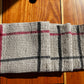 A Classic Linen Dishcloth from Halcyon Yarn, featuring a four-shaft weaving pattern with black and red stripes on beige fabric, rests folded on a wooden surface under warm sunlight.