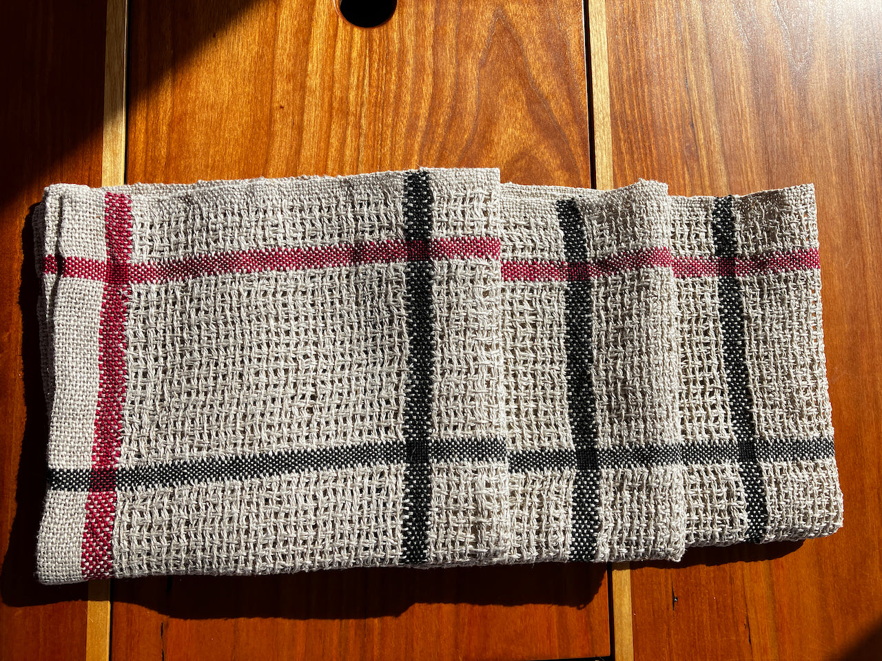 A Classic Linen Dishcloth from Halcyon Yarn, featuring a four-shaft weaving pattern with black and red stripes on beige fabric, rests folded on a wooden surface under warm sunlight.