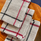 Three Classic Linen Dishcloths from Halcyon Yarn, featuring a Four-Shaft Weaving Pattern with red and black lines on a textured beige fabric, are neatly stacked on a wooden surface.