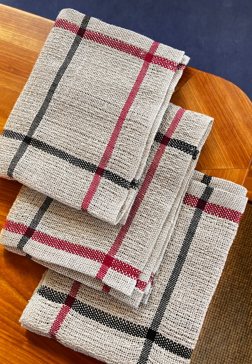 Three Classic Linen Dishcloths from Halcyon Yarn, featuring a Four-Shaft Weaving Pattern with red and black lines on a textured beige fabric, are neatly stacked on a wooden surface.