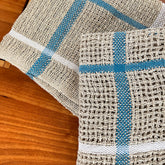 Two Classic Linen Dishcloths from Halcyon Yarn, in beige with blue and white stripes, rest on a wooden surface. With their four-shaft weaving pattern, they exhibit a rustic texture reminiscent of woven mats. Their design brings an absorbent charm to your kitchen decor.