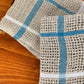 Two Classic Linen Dishcloths from Halcyon Yarn, in beige with blue and white stripes, rest on a wooden surface. With their four-shaft weaving pattern, they exhibit a rustic texture reminiscent of woven mats. Their design brings an absorbent charm to your kitchen decor.