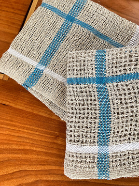 Two Classic Linen Dishcloths from Halcyon Yarn, in beige with blue and white stripes, rest on a wooden surface. With their four-shaft weaving pattern, they exhibit a rustic texture reminiscent of woven mats. Their design brings an absorbent charm to your kitchen decor.