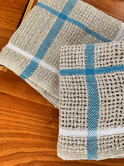 Two pieces from the Classic Woven Linen Dish Cloth Kit by Halcyon Yarn, featuring a beige woven mesh with blue and white stripes, are placed on a wooden surface. These rustic kitchen accessories have an open canvas weave that forms a charming grid pattern, adding texture to any setting.