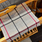 A classic linen dishcloth from Halcyon Yarn is displayed on a wooden loom, featuring a four-shaft weaving pattern with black and red stripes set against a beige backdrop. A shuttle sits atop the loom, surrounded by wooden components and a textured floor that evokes the essence of traditional Irish canvas weave.