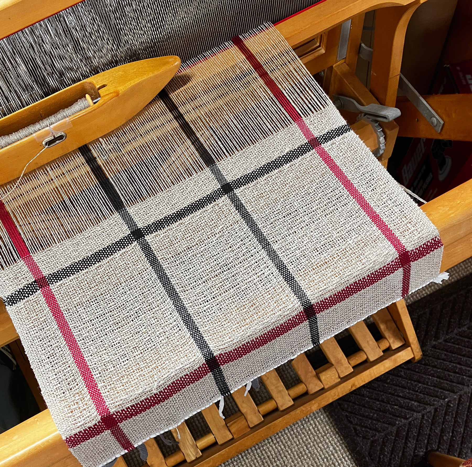 A classic linen dishcloth from Halcyon Yarn is displayed on a wooden loom, featuring a four-shaft weaving pattern with black and red stripes set against a beige backdrop. A shuttle sits atop the loom, surrounded by wooden components and a textured floor that evokes the essence of traditional Irish canvas weave.