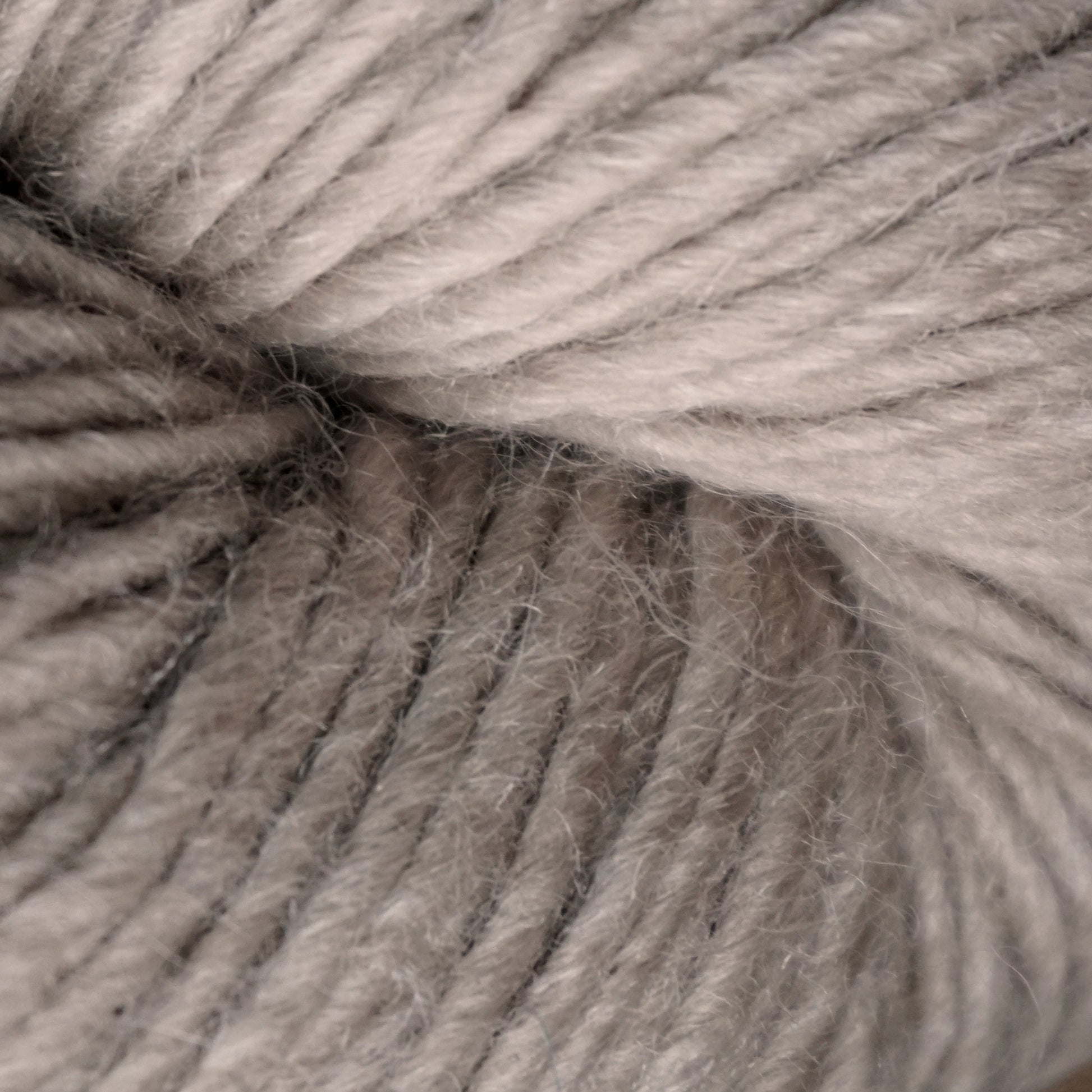 Close-up image of soft, light grey Moonshine by Juniper Moon Farm yarn. The tightly wound fibers create a spiral pattern, showcasing the alpaca yarn's smooth texture and slight fuzziness. The soft, neutral color is consistent throughout, making this Knitting Fever / Euro yarn appear cozy and inviting.