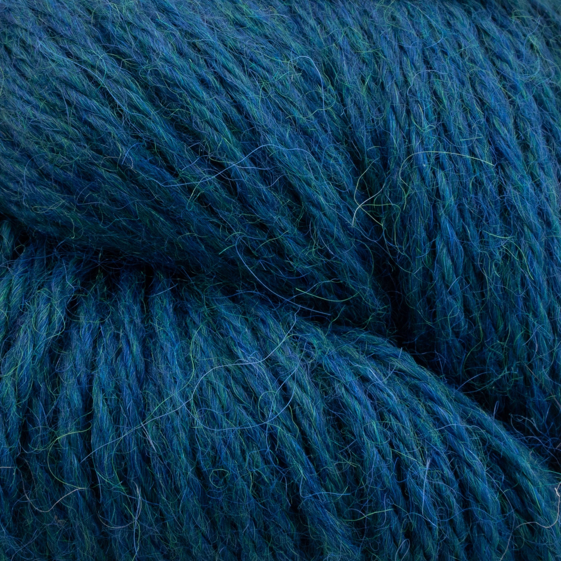 Close-up image of a ball of thick, blue-green Juniper Moon Farm Herriot Baby Alpaca yarn by Knitting Fever / Euro, with visible fibers and threads intertwined. The texture appears soft and woolly, showcasing a rich mixture of blue and green hues characteristic of extrafine baby alpaca.