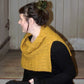 A person with dark hair tied up is seated sideways on wooden steps, gazing to their left. They're wearing a Halcyon Yarn Fortitude Ponchette—a mustard yellow, knitted neck warmer made of Aran weight yarn—over a black long-sleeved top. The background features vertical wooden paneling in a neutral color.