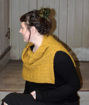 A person with dark hair tied up is seated sideways on wooden steps, gazing to their left. They're wearing a Halcyon Yarn Fortitude Ponchette—a mustard yellow, knitted neck warmer made of Aran weight yarn—over a black long-sleeved top. The background features vertical wooden paneling in a neutral color.