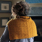 A person with curly hair in a messy bun is shown from behind, wearing the Fortitude Ponchette from Halcyon Yarn. They appear to be in a cozy room with framed pictures on the wall and a stone fireplace in the background. This knitted, mustard-colored ponchette, crafted with Aran weight yarn, makes an ideal winter accessory.