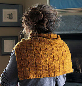 A person with curly hair in a messy bun is shown from behind, wearing the Fortitude Ponchette from Halcyon Yarn. They appear to be in a cozy room with framed pictures on the wall and a stone fireplace in the background. This knitted, mustard-colored ponchette, crafted with Aran weight yarn, makes an ideal winter accessory.