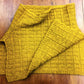 A mustard yellow, hand-knitted rectangular piece of fabric with a ribbed texture, displayed on a wooden surface. The Fortitude Ponchette by Halcyon Yarn is made from Aran weight yarn, making it extremely soft and cozy—perfect as a winter accessory like a scarf or even part of a ponchette.