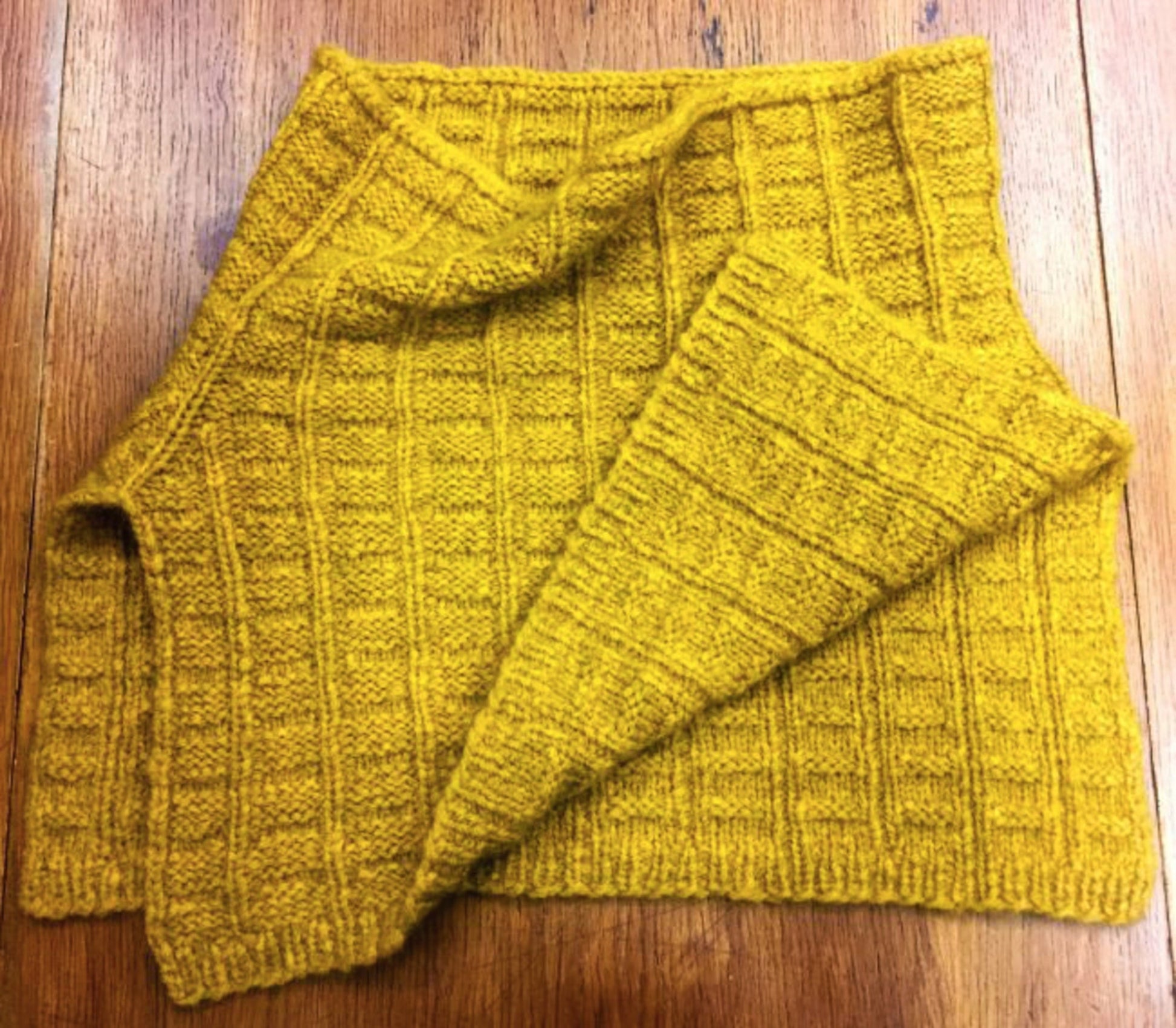 A mustard yellow, hand-knitted rectangular piece of fabric with a ribbed texture, displayed on a wooden surface. The Fortitude Ponchette by Halcyon Yarn is made from Aran weight yarn, making it extremely soft and cozy—perfect as a winter accessory like a scarf or even part of a ponchette.