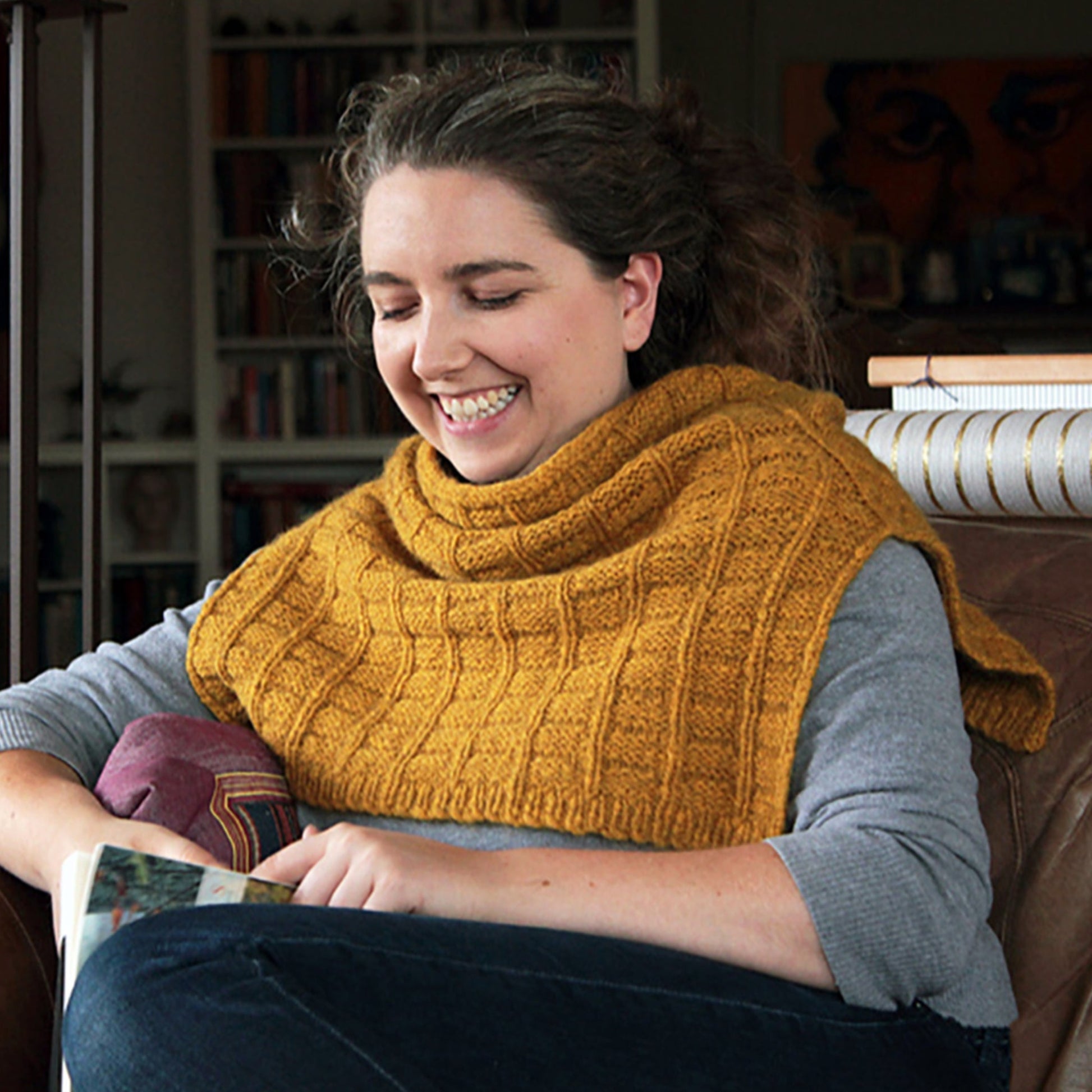 A person with long, curly hair is sitting on a brown leather armchair, smiling while reading. They are wearing a gray long-sleeve shirt and the mustard-yellow Fortitude Ponchette by Halcyon Yarn, crafted from Aran weight yarn and ideal as a winter accessory. The background shows a bookshelf and some artwork on the wall.