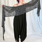 A person with purple hair and tattoos on their left arm stands on a white backdrop, holding up a wide, dark gray knitted All Through The Night Wrap from Halcyon Yarn. They are wearing a sleeveless black dress with loose, baggy pants and black flat shoes.