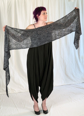 A person with purple hair and tattoos on their left arm stands on a white backdrop, holding up a wide, dark gray knitted All Through The Night Wrap from Halcyon Yarn. They are wearing a sleeveless black dress with loose, baggy pants and black flat shoes.