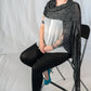 A person with short, purple hair sits on a black folding chair against a plain white background. They are wearing a white button-up shirt, black leggings, teal high heels, and an All Through The Night Wrap from Halcyon Yarn draped over their shoulders. Their arms display several visible tattoos.