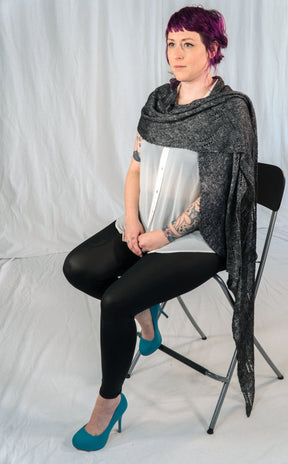 A person with short, purple hair sits on a black folding chair against a plain white background. They are wearing a white button-up shirt, black leggings, teal high heels, and an All Through The Night Wrap from Halcyon Yarn draped over their shoulders. Their arms display several visible tattoos.