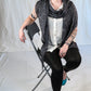 A person with short, purple hair is seated on a folding chair against a plain white backdrop. They are wearing a white shirt, black pants, and turquoise high heels. Wrapped in an All Through The Night Wrap by Halcyon Yarn, made of fingering weight yarn, they have tattoos on their arms and are gazing slightly to the side.