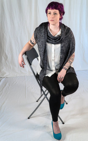 A person with short, purple hair is seated on a folding chair against a plain white backdrop. They are wearing a white shirt, black pants, and turquoise high heels. Wrapped in an All Through The Night Wrap by Halcyon Yarn, made of fingering weight yarn, they have tattoos on their arms and are gazing slightly to the side.