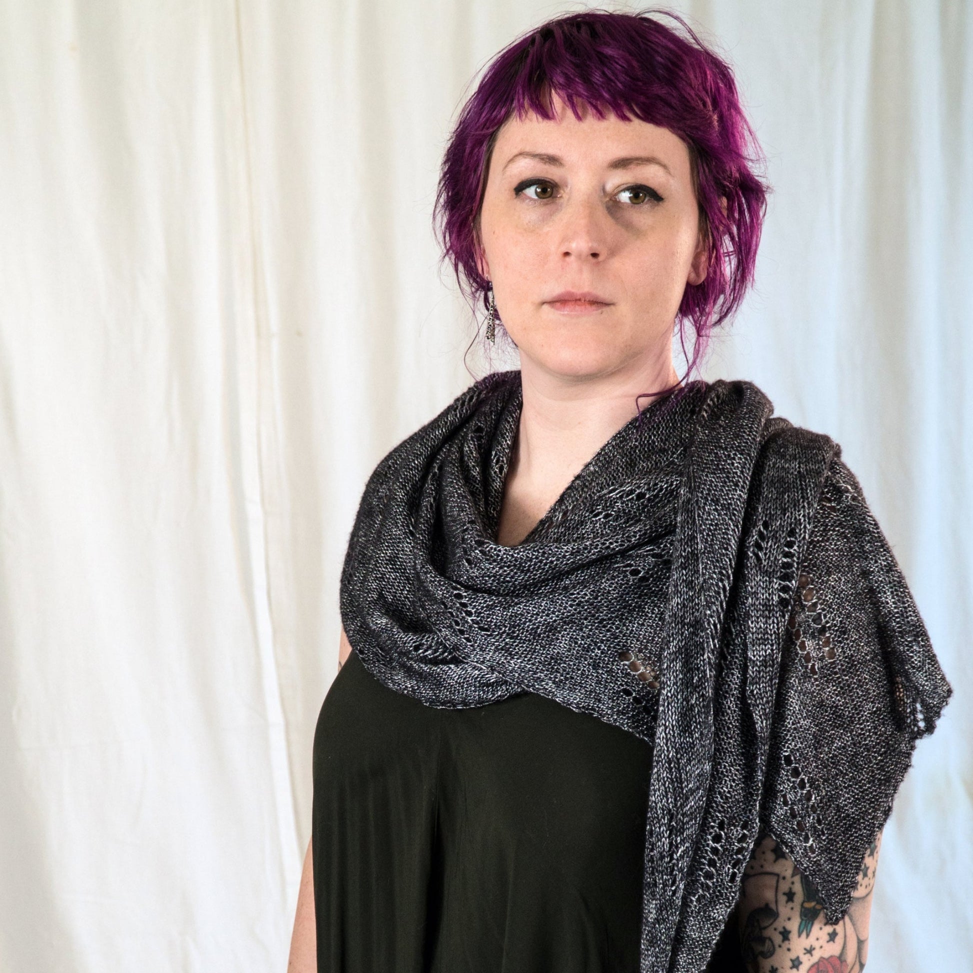 A person with short, vibrant purple hair, dressed in a sleeveless dark top and wrapped in an All Through The Night Wrap from Halcyon Yarn, made from fingering weight yarn, stands in front of a plain white background. They have tattoos on both arms and a neutral facial expression.