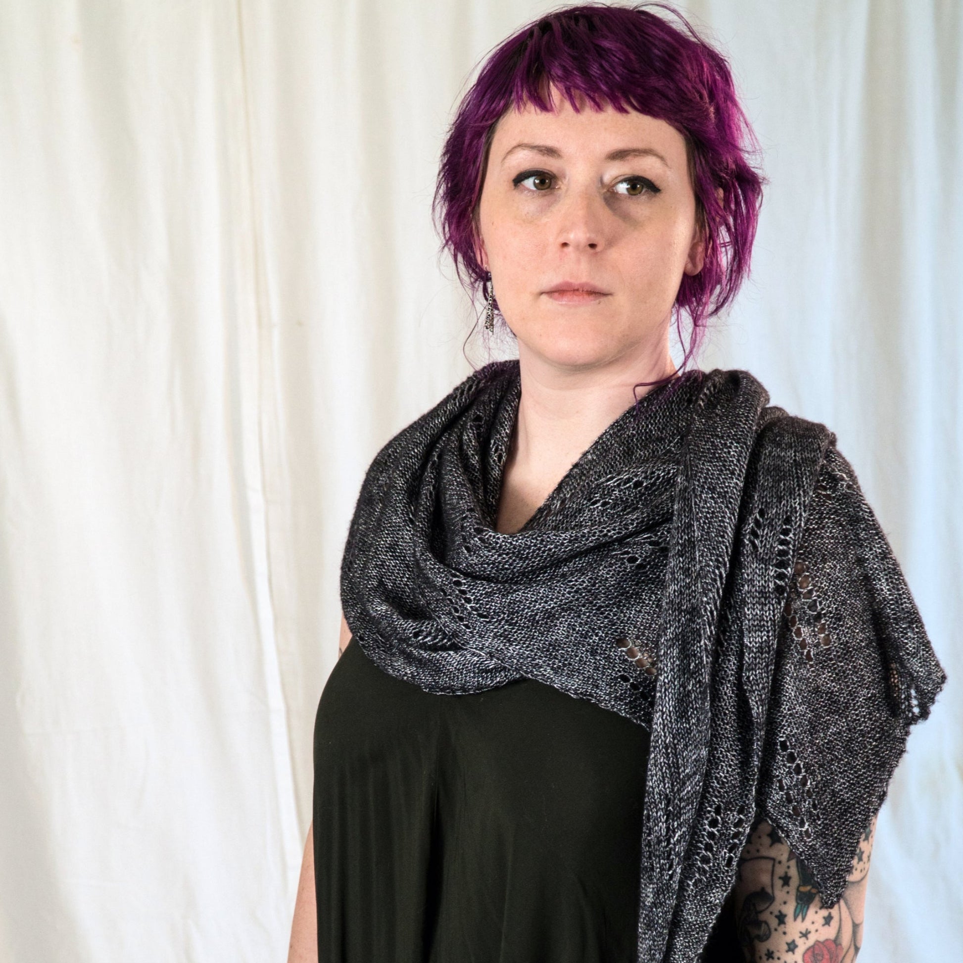 A person with short, vibrant purple hair stands in front of a light-colored curtain, wearing a black top and the All Through The Night Wrap by Halcyon Yarn draped over their shoulders. Made from fingering weight yarn, the large, textured gray shawl exhibits an intricate lace motif. Visible tattoos adorn their right arm as they look to the side.