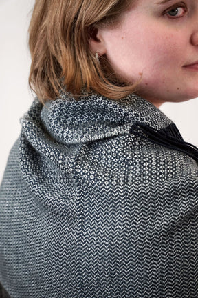 Close-up of a person looking over their shoulder, showing the side of their face and shoulder. They have short, light-brown hair and are wearing the Dancing On The Ceiling woven shawl from Halcyon Yarn, a textured dark gray and white hooded garment with a small twill pattern. The background is plain and light-colored.