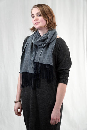 A person stands against a plain background, wearing a dark gray long-sleeve shirt and draped in the Halcyon Yarn's "Dancing On The Ceiling" Woven Shawl, with gray fabric accented by black fringe reminiscent of an intricate weaving project. They have short, light brown hair and are gazing slightly to the side with a neutral expression.