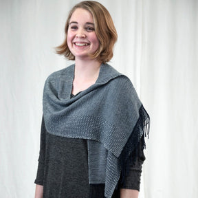 A person is smiling while standing in front of a light-colored curtain. They are wearing a dark gray top paired with Halcyon Yarn's "Dancing On The Ceiling" woven shawl, which is draped over their shoulders. The shawl, from their recent weaving project, features fringed edges and a subtle twill pattern. They have short, light brown hair.