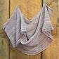 The Pressure Drop Infinity Scarf by Halcyon Yarn is draped on a wooden surface. This light purple knitted scarf features a delicate, airy pattern with decorative open loops along the edges, showcasing its lacy appearance. The simple yet elegant design highlights the craftsmanship of the knitting and the beauty of lace weight yarn.