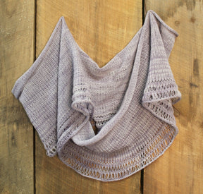 The Pressure Drop Infinity Scarf by Halcyon Yarn is draped on a wooden surface. This light purple knitted scarf features a delicate, airy pattern with decorative open loops along the edges, showcasing its lacy appearance. The simple yet elegant design highlights the craftsmanship of the knitting and the beauty of lace weight yarn.