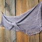 A Pressure Drop Infinity Scarf by Halcyon Yarn, in soft purple yarn with a delicate dropped stitch pattern, hangs draped against a wooden wall. The wood's natural grain and varied tones create a rustic backdrop, contrasting with the scarf's light and cozy texture.