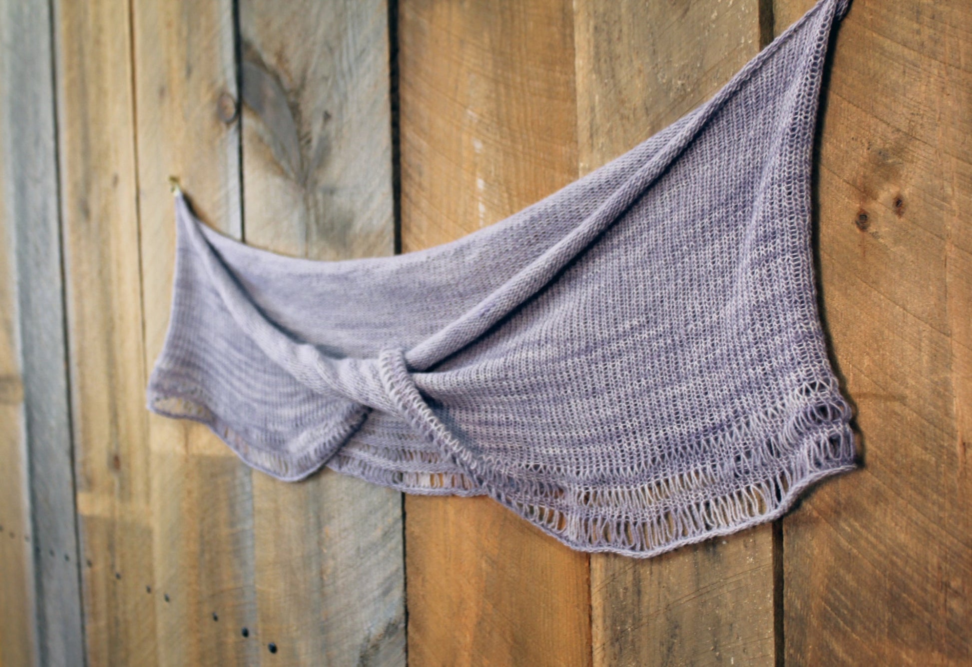 A Pressure Drop Infinity Scarf by Halcyon Yarn, in soft purple yarn with a delicate dropped stitch pattern, hangs draped against a wooden wall. The wood's natural grain and varied tones create a rustic backdrop, contrasting with the scarf's light and cozy texture.