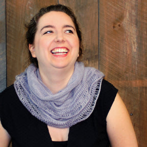 A woman with light skin and dark hair tied back is wearing a sleeveless black top and the Pressure Drop Infinity Scarf, made from lace weight yarn by Halcyon Yarn. She is looking slightly upward and to the side while smiling broadly, with wooden paneling in the background.