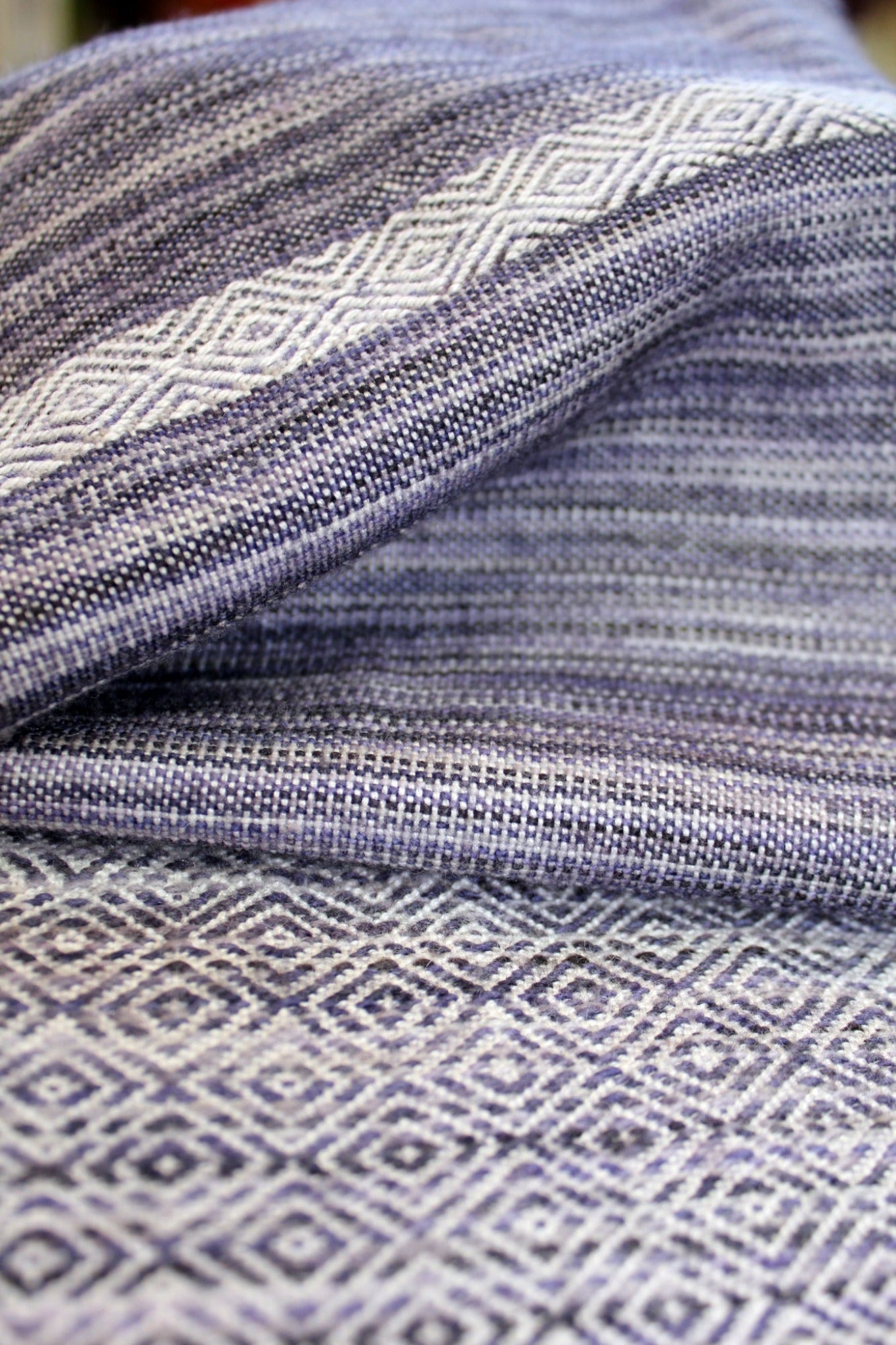 A close-up view of the Serendipity Woven Scarf by Halcyon Yarn, showcasing its intricately woven fabric with a geometric diamond pattern in shades of purple and white. The draped scarf reveals textured details and the precise alignment of the design elements.