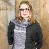 A person with shoulder-length blond hair and glasses stands against a wooden wall, wearing a dark gray top and the Halcyon Yarn Serendipity Woven Scarf in shades of gray and white. They are smiling slightly and looking at the camera.