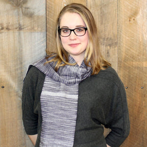 A woman with light skin and straight blonde hair, wearing black glasses, a dark gray sweater, and the Serendipity Woven Scarf by Halcyon Yarn draped around her neck, stands in front of a wooden wall. She has a slight smile and looks directly at the camera.