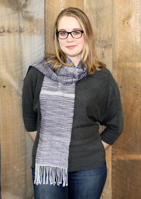 A woman with blonde hair and glasses is standing in front of a wooden wall. She is wearing a grey long-sleeved top, blue jeans, and the Serendipity Woven Scarf from Halcyon Yarn, featuring grey and white hues with fringes made from soft Malabrigo Lace. Her hands are tucked behind her back, and she is smiling softly.