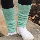 A person wearing Pipeline Legwarmers in mint green, crafted by Halcyon Yarn from Malabrigo Sock yarn, and white canvas sneakers stands on a stone step. The legwarmers reach just below the knee, paired with black leggings. The background features a stone pathway and dirt scattered with leaves.