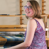 A smiling person with shoulder-length hair and glasses is seated at a weaving loom, wearing the Flower In The Sun Tank Top by Halcyon Yarn. The adjustable straps of the sleeveless top are visible as they face the camera, with their weaving project in progress on the loom in front of them.