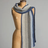 A vintage dress form displays a crocheted scarf draped around its shoulders. Crafted using a free pattern from Yarn & Soul, the Sideways Scarf features vertical stripes in shades of blue and gray, with the visible crochet stitches giving it a textured appearance. The background is plain gray.