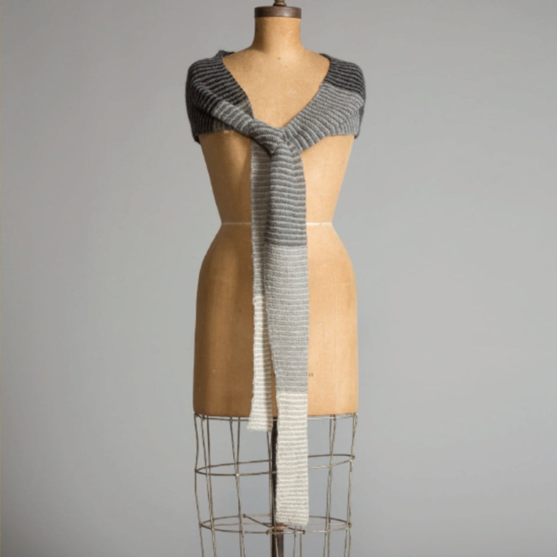 A dress form adorned in a Crescent Wrap by Yarn & Soul, elegantly draped around the shoulders and cascading down in front. The knit wrap transitions from dark to light gray towards the middle and bottom sections. The background is plain and neutral in color.