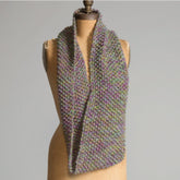 A brown mannequin showcases the Five Strand Cowl draped around its neck, featuring a blend of green, purple, and beige hues. Made with Superfine 400 Alpaca Braids, this textured multicolored knitted scarf from Yarn & Soul highlights intricate knitting patterns against a plain grey background.